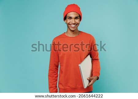Young smiling happy fun african american man 20s in orange shirt hat hold closed laptop pc computer look camera isolated on plain pastel light blue background studio portrait People lifestyle concept