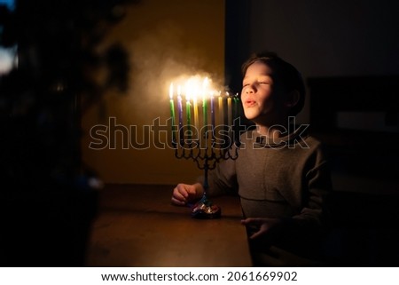 The child blows out the menorah for Hanukkah on the windowsill. The boy in the kippah sitting by the window. Jewish holiday. Tradition is a religious ritual. Sunset. The first star. Judaism holy