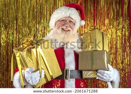 Santa Claus on golden foil backdrop. Emotional senior male model old man with a natural white beard in the role of Father Christmas bringing wrapped magic boxes Xmas gifts.
