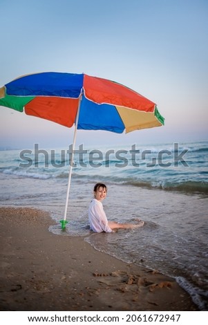 European child sits on the beach by the sea under a rainbow umbrella from the sun