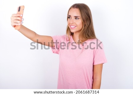 Isolated shot of pleased cheerful Young Caucasian girl wearing pink T-shirt on white background, makes selfie with mobile phone. People, technology and leisure concept