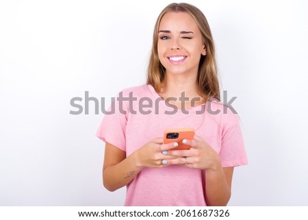 Pleased Young Caucasian girl wearing pink T-shirt on white background using self phone and looking and winking at the camera. Flirt and coquettish concept.