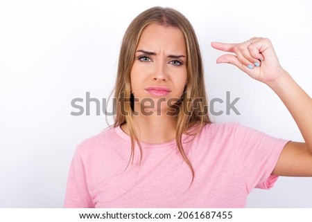 Upset Young Caucasian girl wearing pink T-shirt on white background shapes little gesture with hand demonstrates something very tiny small size. Not very much