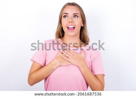 Happy smiling Young Caucasian girl wearing pink T-shirt on white background has hands on chest near heart. Human emotions, real feelings and facial expression concept.