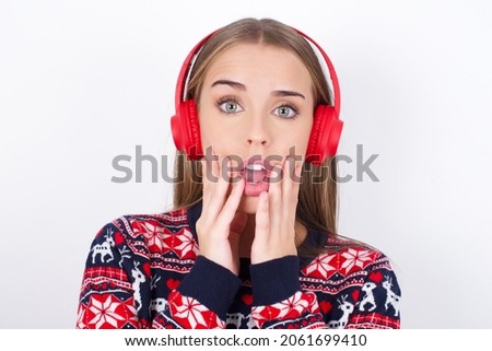 Shocked Young caucasian girl wearing christmas sweaters on white background stares fearful at camera keeps mouth widely opened wears wireless stereo headphones on ears