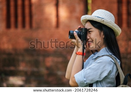 Young asian backpacker female wearing hat smiling while has trip in historic site, she use camera take a photo with happy