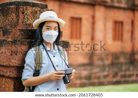 Young asian backpacker female wearing hat and protection mask while traveling in historic site, she use camera take a photo with happy
