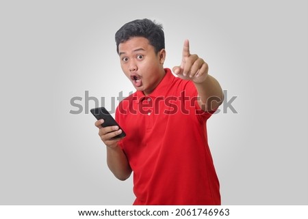 Portrait of shocked Asian man in red polo shirt standing against gray background, looking and pointing forward with his finger while using mobile phone. Wow face expression