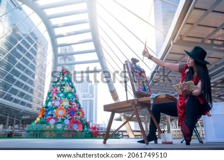 Female street artist in a hat paints a picture on the town square. Christmas tree background, Christmas holiday