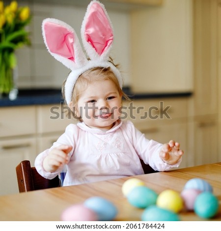 Cute little toddler girl wearing Easter bunny ears playing with colored pastel eggs. Happy baby child unpacking gifts. Adorable healthy smiling kid in pink clothes enjoying family holiday