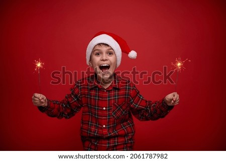 Christmas and New Year celebration concept for advertisement. Funny amazing handsome child boy in Santa hat with sparklers against red colored background with copy space