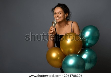 Attractive woman smiles looking at camera, poses with glass of sparkling wine and luxury festive shiny golden and green metallic air balloons against gray wall background with copy space
