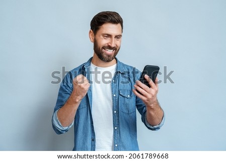 Happy satisfied man looking at mobile phone screen gesturing yes with clenched fist isolated on blue studio copy space. Overjoyed excited joyful guy make winner gesture read message about reward Royalty-Free Stock Photo #2061789668