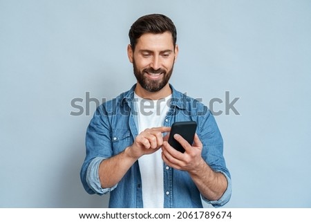 Smiling caucasian man using smartphone modern device looking at mobile screen scrolling enjoying communication with friends. Handsome guy standing isolated on blue background studio copy space Royalty-Free Stock Photo #2061789674