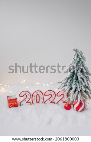 Happy new year 2022 red and white numbers under a fir tree in snow with warm bokeh.  Christmas and festive concept. Holiday greeting card design. White background. Free copy space. Vertical picture