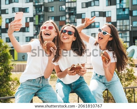 Three young beautiful smiling hipster female in trendy summer  clothes.Sexy carefree women posing in the street.Positive models taking selfie in sunglasses.Holding juicy burger and eating hamburger Royalty-Free Stock Photo #2061810767
