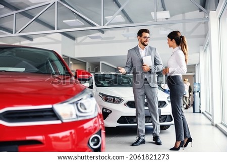 Smiling car seller standing in car salon with customer and showing around cars on sale. Royalty-Free Stock Photo #2061821273