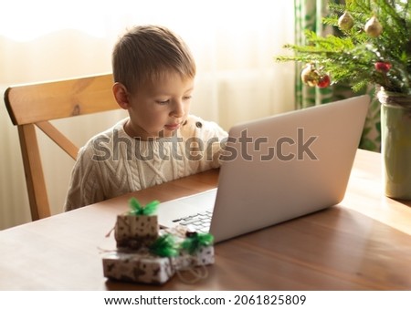  Little boy in a white sweater sits at a laptop and writes a letter to Santa Claus.