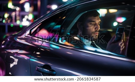 Handsome Businessman in a Suit Commuting from Office in a Backseat of His Car at Night. Entrepreneur Using Smartphone while in Transfer Taxi in Urban City Street with Working Neon Signs.