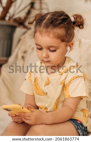 A three-year-old girl with headphones using a phone at home on the couch,playing games and watching cartoons.Children's leisure, technologies, gadgets and children concept.Selective focus.