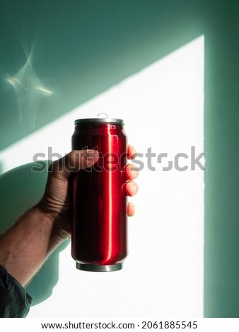 Red thermo can bottle in male hands blue turquoise wall background harsh shadow. Hot tea coffee beverages drinking in cold autumn winter weather. Durable stainless steel reusable eco-friendly thermos.