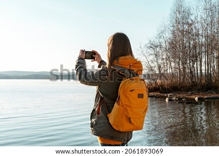 Rear view of young blonde woman with yellow backpack taking photo on smartphone of autumn landscape lake, using modern technology beauty in nature, fall season walking photographing