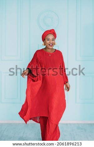 Beautiful positive african american woman wearing red ethnic clothes and headscarf, walking on camera with happy face smiling. Studio shot of pretty lady on blue background