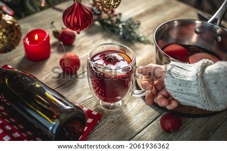 Woman in a sweater is holding a clear mug with fragrant spicy mulled wine decorated with fruits and spices. Concept of a cozy festive atmosphere, New Year and Christmas mood.