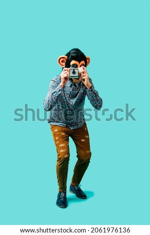 man, wearing a monkey mask, is taking a picture with a retro instant camera, standing on a blue background