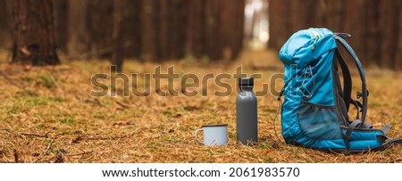 Backpack, thermos and travel mug in forest. Hiking equipment on footpath in woodland. Panoramic view