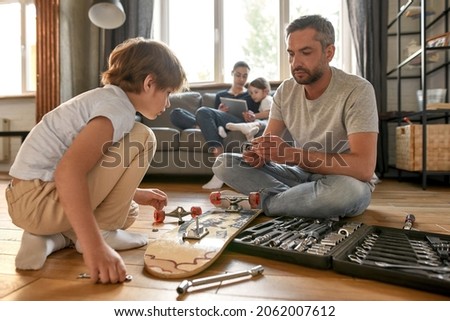 Caucasian dad and teenage son sit on floor at home repair skateboard with toolset in box. Young father teach small teen boy child fixing board with tools equipment. Handyman, fatherhood concept. Royalty-Free Stock Photo #2062007612