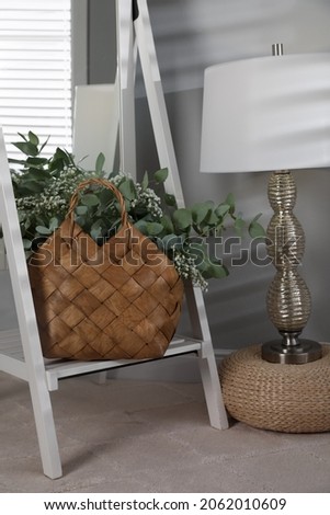 Stylish straw bag with beautiful bouquet and lamp indoors