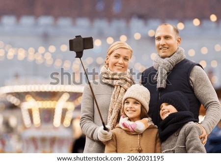 winter holidays, technology and people concept - happy family taking picture with smartphone and selfie stick over christmas market or amusement park background