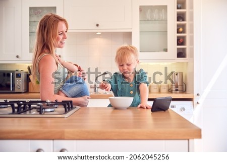 Girl At Kitchen Counter Eating Breakfast Watching Mobile Phone Whilst Mother Feeds Baby