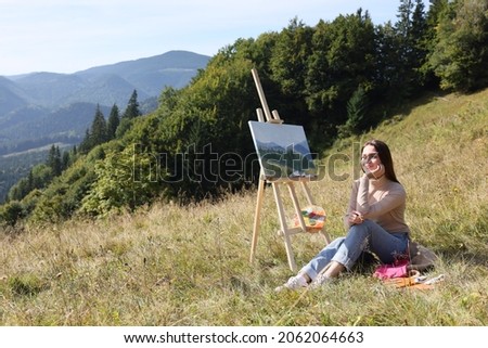 Young woman near easel with drawing in mountains, space for text