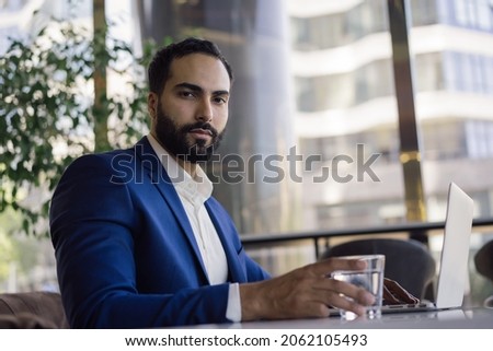 Handsome smiling arabic businessman using laptop computer planning start up in modern office. Confident middle eastern manager wearing suit holding glass of water working online sitting at workplace