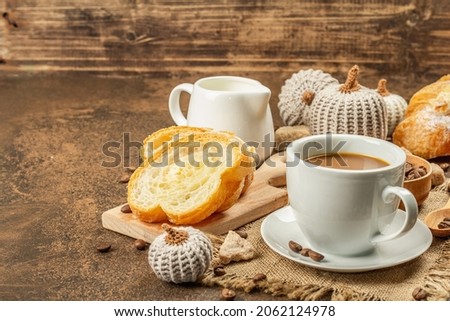 Breakfast concept with a cup of coffee, croissants, milk jug, and decorative crochet pumpkins. Warm brown stone concrete background, copy space