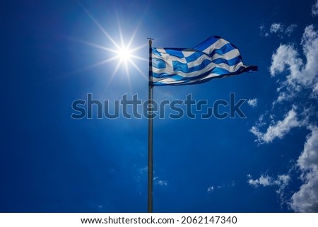 The national flag of Greece is waving in the clear blue Greek sky. The white cross symbolises Eastern Orthodox Christianity, the prevailing religion of Greece. Royalty-Free Stock Photo #2062147340