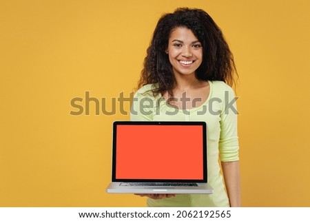 Smiling jubilant cheerful african american young brunette woman 20s wears green shirt hold use work on laptop pc computer with blank screen workspace area isolated on yellow background studio portrait
