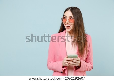 Young fun surprised excited woman 20s in pastel pink clothes glasses using mobile cell phone chatting online look aside isolated on blue background studio portrait. People lifestyle technology concept