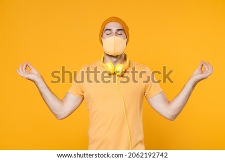 Young man in t-shirt headphones face mask safe from coronavirus virus covid-19 during pandemic quarantine hold hands in yoga gesture, relaxing meditating isolated on yellow background studio portrait