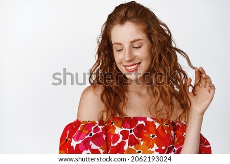 Beautiful young redhead woman playing with her curly hair, looking down with flirty coquettish smile, standing against white background