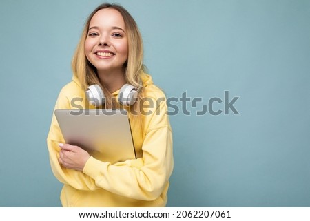 Photo portrait of beautiful attractive positive happy smiling blonde young woman with sincere emotions wearing hipster stylish yellow hoodie isolated over blue background with copy space, holding