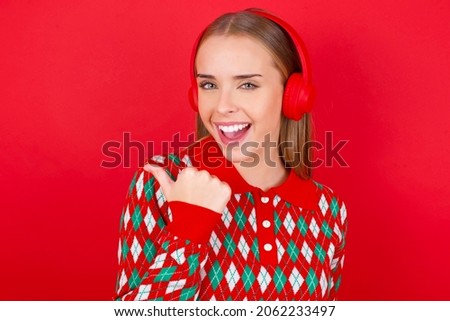 Young caucasian girl wearing Christmas sweater on red background listens audio track via wireless headphones points thumb away advertises copy space