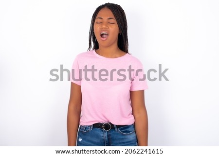 Young girl with dreadlocks wearing pink t-shirt on white backgrond yawns with opened mouth stands. Daily morning routine