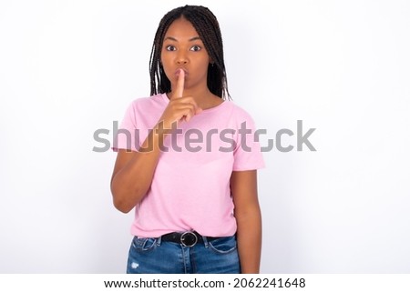 Surprised Young girl with dreadlocks wearing pink t-shirt on white backgrond makes silence gesture, keeps finger over lips and looks mysterious at camera