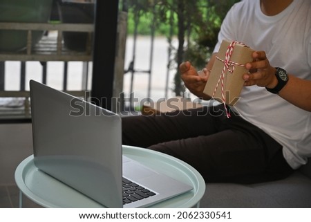 Cropped image of a young man showing a present box while making a video call via the computer laptop in the comfortable living room.