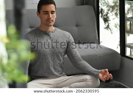 Photo of a young man with wireless earphones doing yoga and meditating on the living room floor over the comfortable sofa as a background. Royalty-Free Stock Photo #2062330595