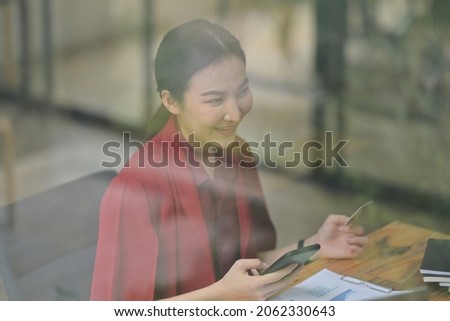 Photo through the window of a beautiful woman holding a credit card and smartphone at the wooden working desk.