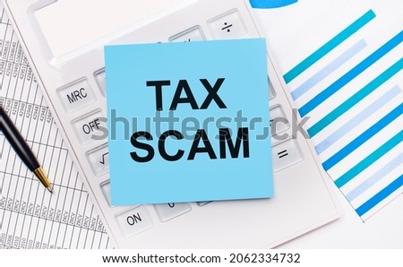 On the desktop is a white calculator with a blue sticker with the text TAX SCAM, a pen and blue reports. Business concept